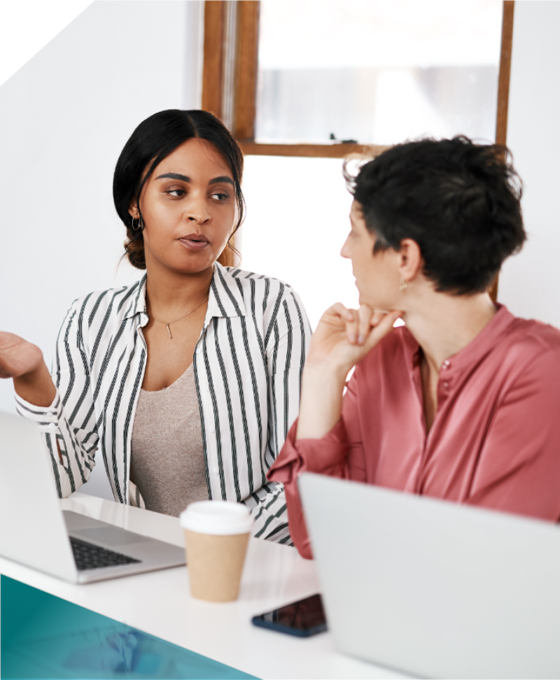 Two young professional women sit beside each other in an office with laptops having a discussion